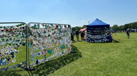 Shariqua Ahmed/BBC Grilled frame with thousands of paper hearts placed on green grass - and a blue tent with more rows of paper hearts displayed on it.