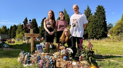 Vlad's family, Mia, Masha, Anna & Graham at his graveside in Southampton which is covered in tributes and marked by a home made wooden cross with his name on