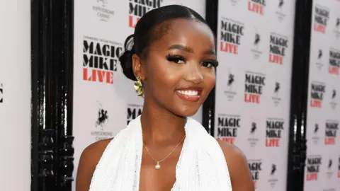 Getty Images Diamanté Laiva at an event. She wears a low-cut white halter-neck dress with a silver necklace. Her dark hair is tied back in a low bun and she has a glam make-up look. She smiles at the camera, her chin tilted down towards her left shoulder. 