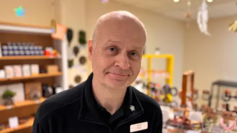 A bald man wearing a black polo shirt, with a name badge saying 'Paul', stands in a shop with merchandise behind him. 