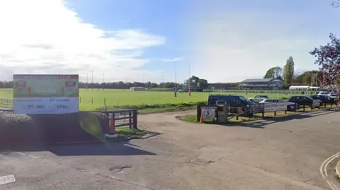 A view of the entrance to the Cambridge Rugby Football Club. A large sports field can be seen just beyond a small car park, in which cars are parked. 