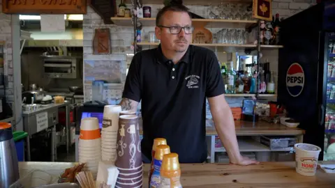 BBC News/Eloise Alanna Chris Scott stands behind the counter at his Whistle Stop cafe. He is wearing a collared black t-shirt with his cafe's logo in white. In front of him are disposable coffee cups and three bottles of coffee creamer. Behind him are bar shelves with alcohol bottles and drinking glasses. He is wearing square-framed seeing glasses, has a moustache and goatee and a short haircut, and is looking off to the right.