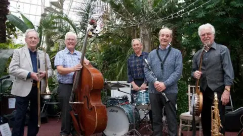 Courtesy of The Capstone Theatre Jazz band The Savoy Jazzmen standing with their instruments smiling in an atrium.