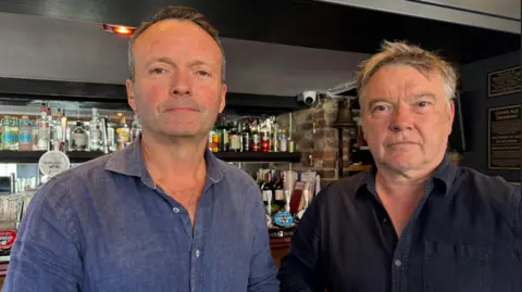 Adrian Rothera and Rory Fry-Stone, co-owners of Pilgrim brewery which runs the Cage Wine Bar in Reigate. Adrian, on the left, is wearing a blue shirt and has greying hair. Rory, who also has greying hair, is wearing a navy shirt.