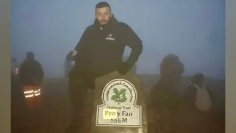 Lee, wearing a black coat, at the summit of Pen y Fan. He has his hand on a stone marker which marks the mountain's height at the top