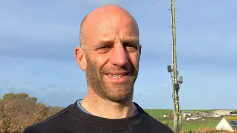 Bald headed man with a short well clipped beard and moustache looking at the camera. There is a very cluttered telegraph pole next to him in the distance.
