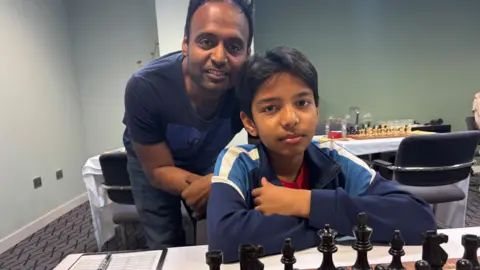 Simon Thake A young boy sits in front of a chess board with his arms folded. An older man, possibly his father, looks over his shoulder.