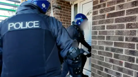 Police in riot gear bash down a white door