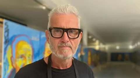 A male artist with short grey hair, who is wearing thick black framed glasses and a black T-shirt. He is standing in a room which has his 50 metre long painting running along the walls.
