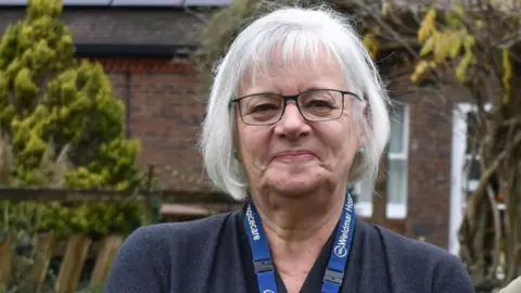 Weldmar Hospicecare Caroline Hamblett smiles at the camera in a garden at the hospice. She has short, white hair and glasses.