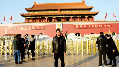 Courtesy Yatai She Zhijiang wearing a black leather jacket and black trousers at Tiananmen Square, in front of yellow barricades 