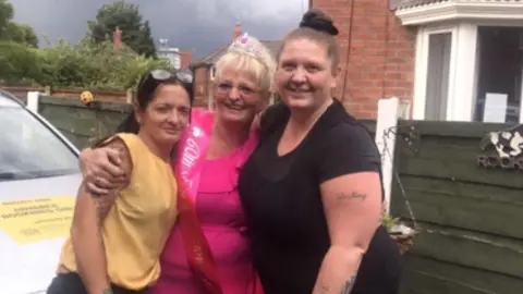 Michelle Maher Three women, two younger and one older, stand hugging. The women and smiling and the woman in the middle (Rita) is wearing a crown and sash reading '60th'.