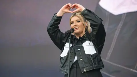 Getty Images Ella Henderson is stood on the stage at BBC Radio 1's Big Weekend 2024, behind a microphone. She is smiling and holding her hands in a heart shape above her head. She wears a black denim jacket with white pockets and black trousers. She has blonde hair and it is tied up.