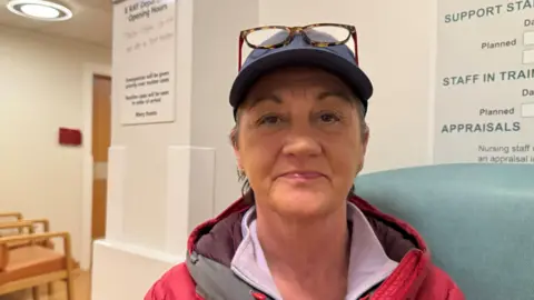 Tracy Waugh is sitting on a green hospital chair and looking directly at the camera with a neutral expression on her face. She is wearing a pink coat, a blue cap with glasses resting on top.
