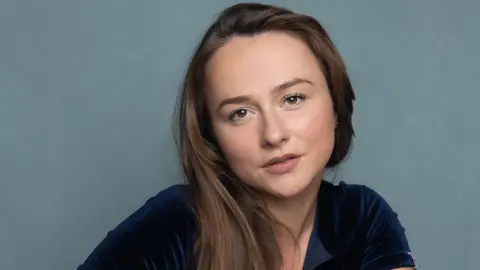 Elizabeth G Photography Headshot of Sheehy. She wears a deep blue velvet jumpsuit. Her brown hair is draped over her right shoulder.
