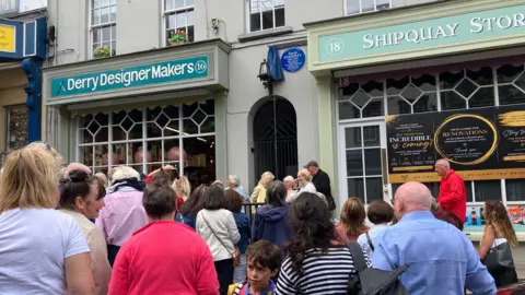 A large group of people are standing on a road in front of two shopfronts. A blue plaque is visible on a wall between the shop fronts.