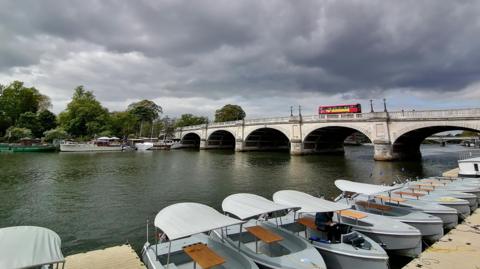A red bus on a bridge over a river, with a row of small boats tied to the riverside in the foreground and dark clouds above