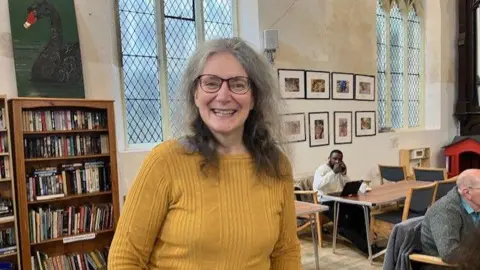 Claire Foullon, who s standing up and smiling inside The Strand Centre. She has just below the should grey and dark hair, she is wearing dark glasses and a mustard yellow knitted top. Behind her are two bookcases, as well as top eople sitting a cafe tables.