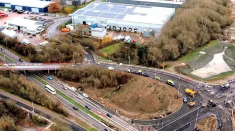 A large junction construction project with construction vehicles working on a flyover near an industrial site.