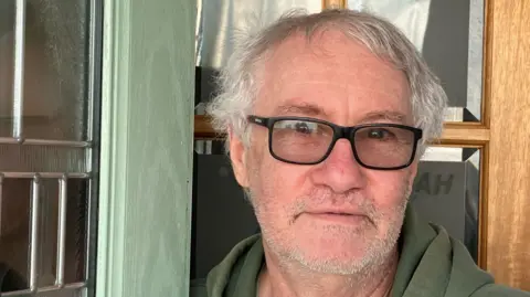 Grace Wood/BBC A man with short grey hair and a short grey beard. He is wearing a green hoodie and black-framed glasses. He is standing behind a green door frame.