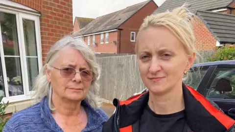 Michelle Watkins and her mother Pam Tovey. They are standing on their front drive. Pam is on the left wearing glasses and a blue jumper. Michelle is wearing earrings and a has a black and red jacket on. 