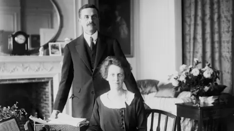 PA Media Waldorf Astor, the 2nd Viscount Astor, and his wife Nancy Astor. Nancy is sitting behind a desk and Waldorf is standing up behind her. The photo is black and white and both are wearing period clothing.