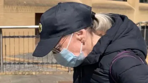 A woman wearing glasses, a dark blue coat, a dark blue baseball cap with a medical mask covering her face. The picture captures the side of her face.