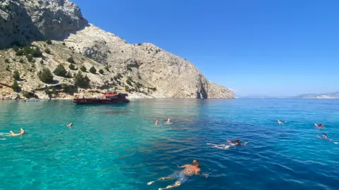 A turquoise sea below a light-coloured cliff face. In the sea, there are several people swimming, spread out across the image. In the background, there is a red, large boat.