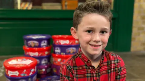 Teddy, a seven-year-old boy with freckles and wearing a red tartan shirt, smiles at the camera. Behind him, slightly out of focus, are stacked round tubs of chocolates, such as Heroes, Quality Street and Celebrations.
