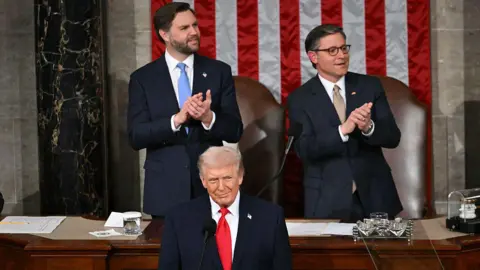Donald Trump stands at a podium as he shares his State of the Union address. Vice-President JD Vance & House Speaker Mike Johnson are seen in the background applauding. 