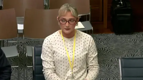 A woman in a white button top, with black polka dots wears red-framed glasses. She sits on a grey leather chair wearing a yellow lanyard around her neck. She has medium length blonde hair. 