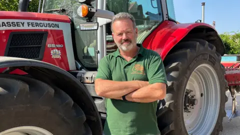 Richard Bramley stands in front of a red tractor, parked in front of a hedgerow. He wears a green polo short with an orange logo on the chest, and has short grey hair and a grey beard.