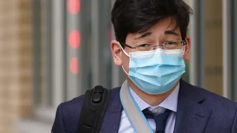 Jonathon Dean wearing a navy suit with a navy tie and a white shirt and glasses. He is walking outside of a court building with the strap of a black rucksack over one shoulder and a facemask on.