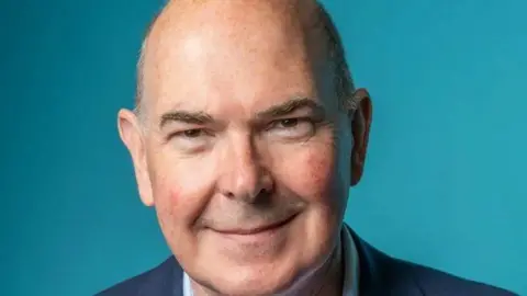 A bald, older man wearing a navy blue suit smiles against a lighter blue background.