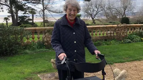 Joy Palfreman is standing in a garden with a walker. She is wearing a navy blue jacket and is smiling at the camera.