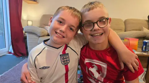 BBC Two young boys with blonde hair are wearing England and Arsenal football t-shirts standing arm in arm. They are in a living room and both smiling at the camera. Josh, on the left, is wearing dark-rimmed glasses. Behind them is a beige-coloured sofa.