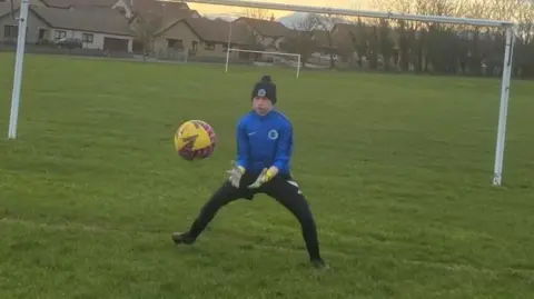 Golebiewski family Adam Golebiewski training as a goalkeeper before his injury, wearing dark trousers, a blue top and a dark hat, preparing to catch a yellow football, among goalposts.