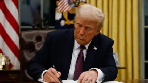 Trump sits at a desk and signs a paper with a pen