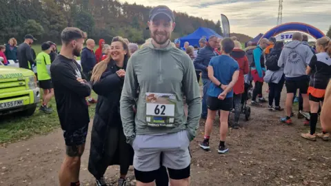 Billy Green Billy Green taking part in an organised run. He has a number 62 on his stomach and there are lots of people around him. 