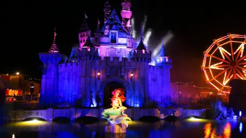 A wide shot of Banksy's Dismaland at night, showing a Disney-like castle lit in purple and a Little Mermaid-like statue