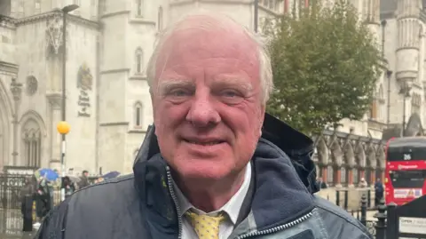 Sir Edward Leigh who has short white hair wearing a black raincoat and white shirt with a yellow tie.