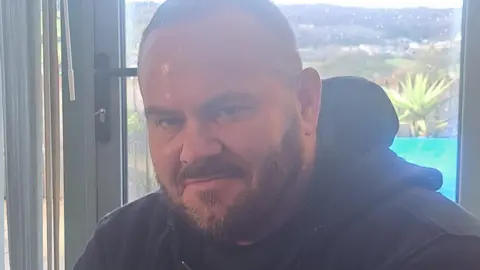 Contributed Allis smiles into the camera. He is bald and wearing a black hoodie. Behind him a florescent yellow paddling pool can be seen from the window and a vista of a valley.