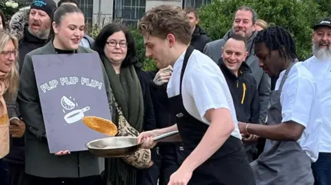 A chef dressed in a white shirt with a black apron flips a pancake ahead of a second chef in a white shirt and a grey apron.