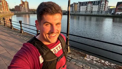 A man with short brown hair wearing a red tshirt and a black running vest, he is smiling at camera as he takes a selfie, running alongside the water in Belfast. There are high rise waterfront apartments visible on the horizon.