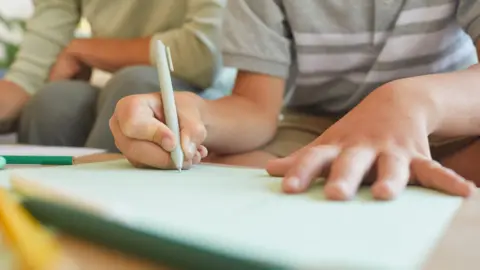 A boy wearing a grey and white t-shirt, his face out of camera shot, writes in an exercise book with a biro, as an adult in grey trousers and a pale cream pullover sits beside him and watches.