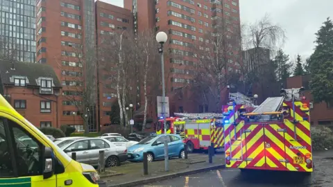 Emergency vehicles in a car park,  including two fire engines and an ambulance gathered outside a block of flats. 