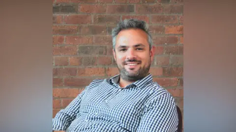 Jon Eaton Jon leaning back in a chair in a mid-shot, smiling to camera and wearing a blue and white checked open-necked shirt. He has a greying beard and hair and there is a brick wall behind him