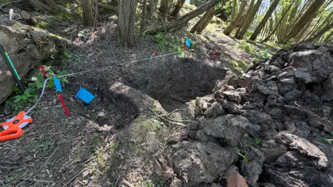 An area of woodland has been cordoned off with blue rope. There are two deep holes side by side in the mud and four orange pegs mark the spots. The orange cordon holders are on the ground and there is a old looking tree in the background. Some parts of the ground are lit by the sun. 