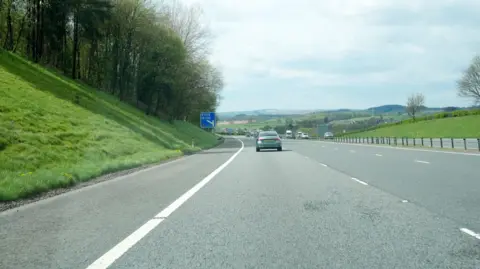 Malcolm Neal A stretch of motorway with a few cars on it and a blue sign in the distance. There are trees up a grass bank next to it and hills on the horizon.
