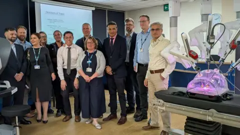 Sheffield Teaching Hospitals The machine looks like a plastic container with purple light inside, with lots of metal rods poking into it. Behind it are 11 people wearing smart clothing, many of them with blue NHS lanyards.